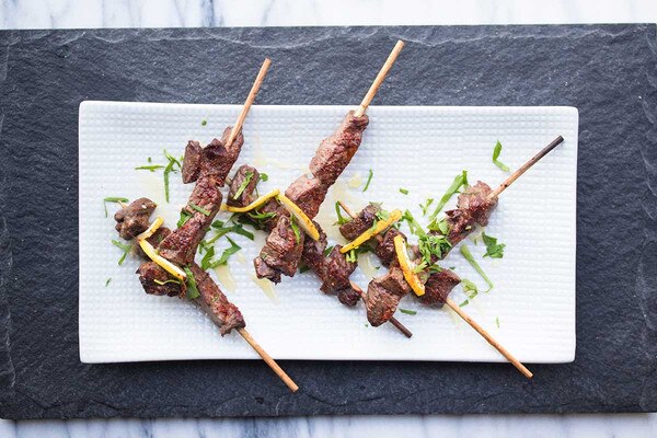 5 skewers of cooked bison meat with lemon, parsley & espelette pepper flakes, white plate with slate charger on marble background