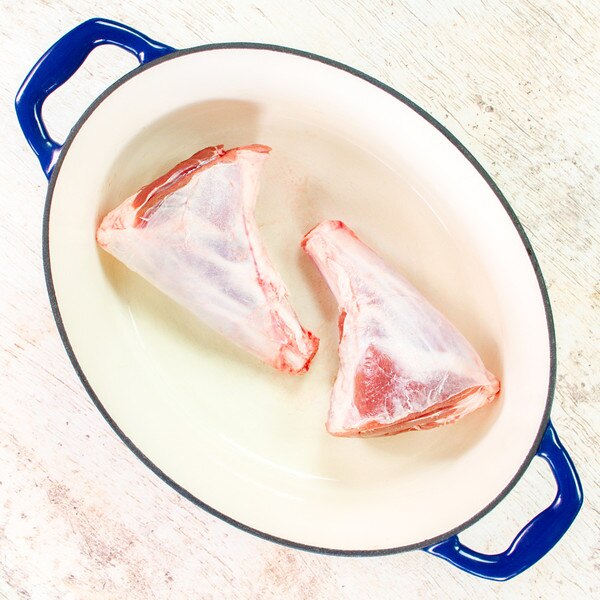 2 raw grass-fed lamb hindshanks in an oval pot with blue rim & handles on whitewashed wood background