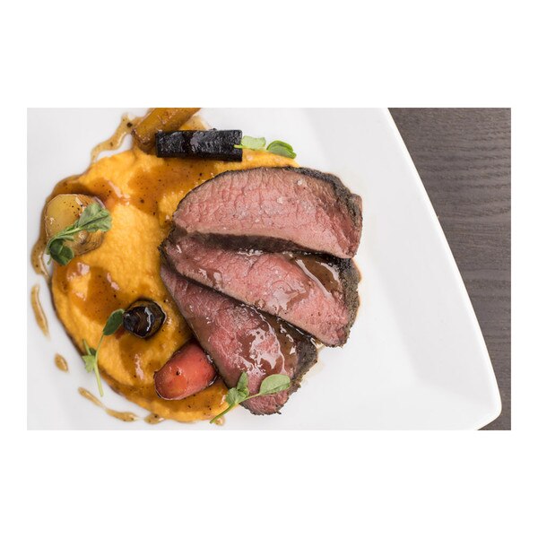 Cooked, sliced Canadian Elk striploin, rainbow carrots, orange carrot cream, allspice gastrique, pea shoots, square white plate, gray concrete background