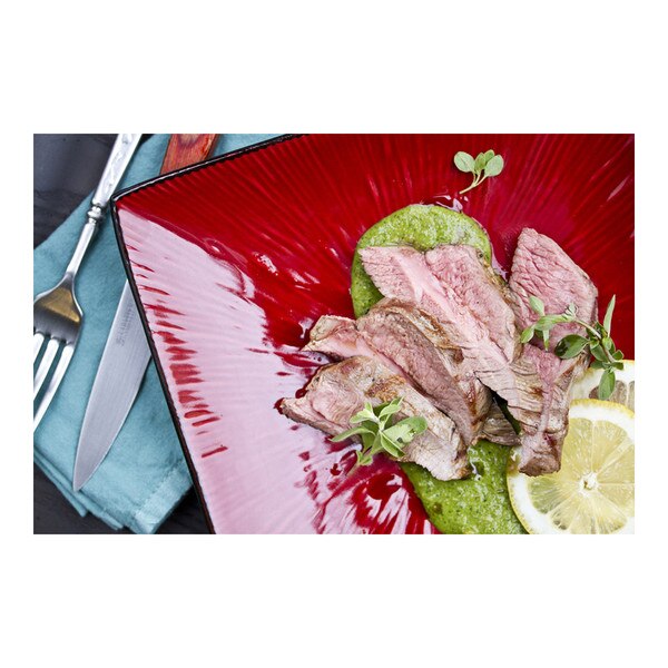 Cooked slices of boneless grass-fed lamb leg with green pesto sauce, lemon slice & fresh herbs, square red plate, blue napkin, fork & knife