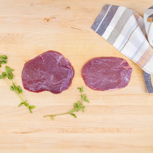 Two raw venison flank steaks with herb sprigs and a striped napkin on a pale wood background.