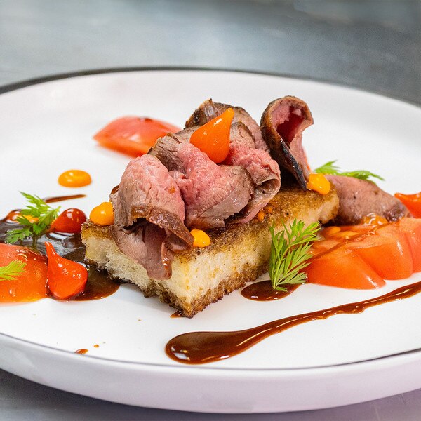 Slices of pan-seared venison flank steak on toasted bread with sliced tomato, tiny teardrop peppers, herb sprigs, dots of orange cheese sauce, and dribbles of brown sauce.