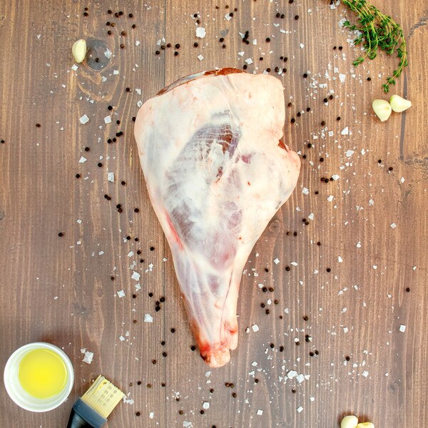 Raw bone-in grass-fed lamb leg, bowl of oil, basting brush, whole garlic cloves, salt & pepper on brown-stained wooden board