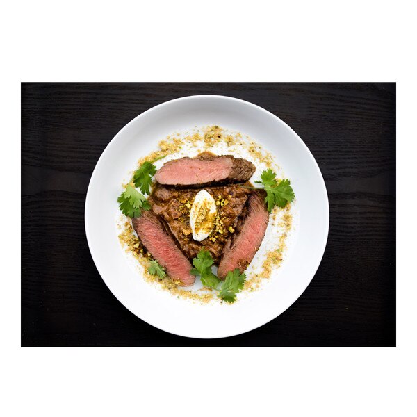 Cooked slices of boneless grass-fed lamb striploin with apricot sauce, chopped pistachios, cilantro sprigs, dollop of yogurt, round white plate on black background