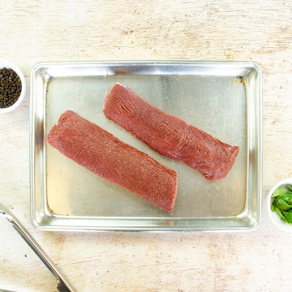 2 raw boneless grass-fed lamb striploins on a metal sheet pan, bowls of peppercorns & mint leaves, tongs, on whitewashed wood background