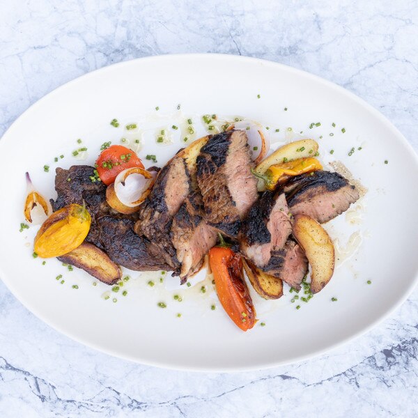 Slices of cooked Ocean Beef boneless ribeye with roasted red and yellow tomatoes, fingerling potatoes, and chopped herbs on an oval plate against a blue-and-white marble background.