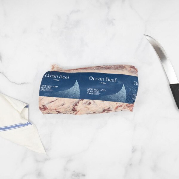 Packaged Ocean Beef boneless ribeye roll with a blue label against a white marble background with a blue-striped towel, left, and a carving knife, right.