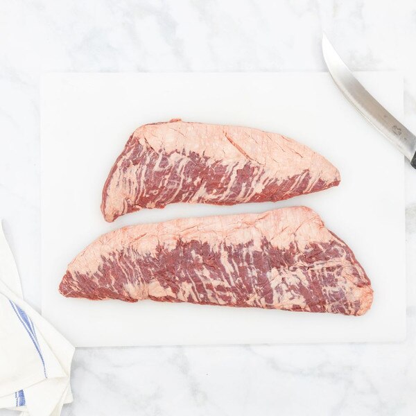 A white cutting board on a gray-streaked white marble background shows two raw Ocean Beef skirt steaks with a blue-striped, white towel in the lower left, and the curved blade of chef’s knife in t
