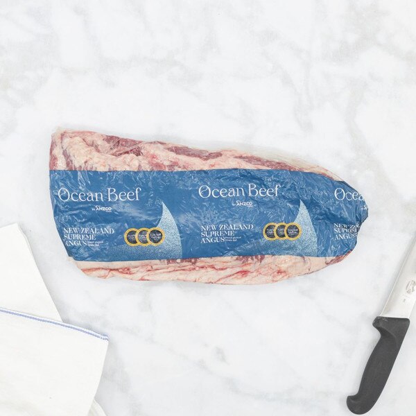 Two vacuum-sealed packs of Ocean Beef New Zealand Supreme Angus skirt steaks on a gray-streaked white marble background with a blue-striped towel on the left, and the black handle of chef’s knife.