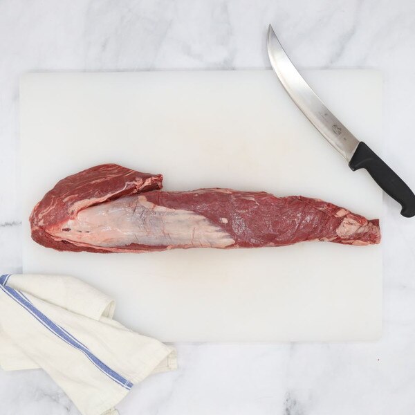 Raw Ocean Beef grain-fed Angus tenderloin on a white cutting board against a white marble background with a blue-striped towel, lower left, and carving knife, upper right.