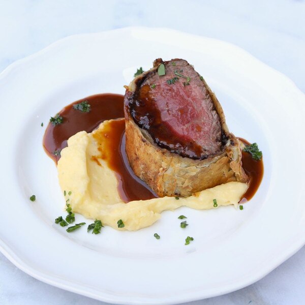 Pastry-wrapped slice of Beef Wellington made with Ocean Beef grain-fed Angus tenderloin atop mashed potatoes with chopped herbs and a brown sauce on a round white plate.