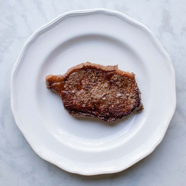 A cooked grain-fortified NY strip strip steak on a round white plate against a white marble background.
