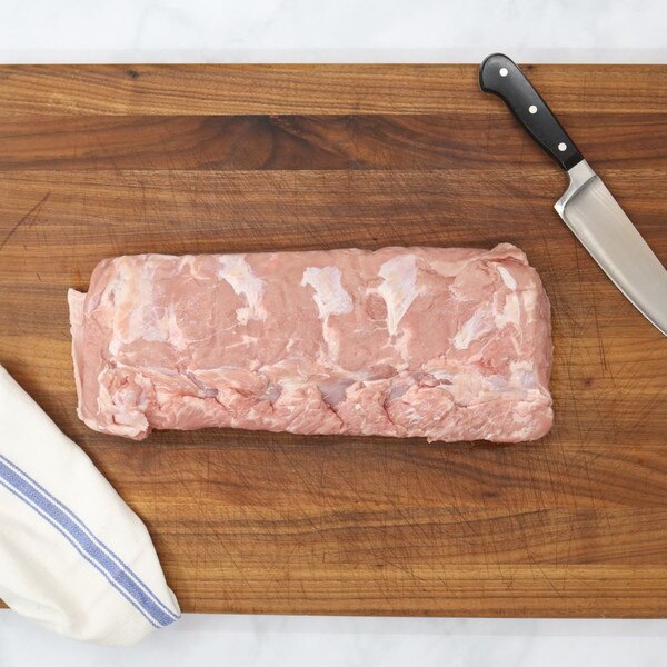 A raw, boneless milk-fed French veal striploin on a wooden board with a blue-striped white towel to the left and a chef’s knife on the right.
