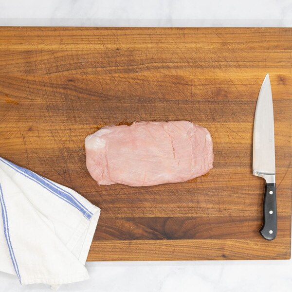A whole, raw flank of milk-fed French veal on a wooden board with a blue-striped white towel to the left and a chef’s knife on the right.