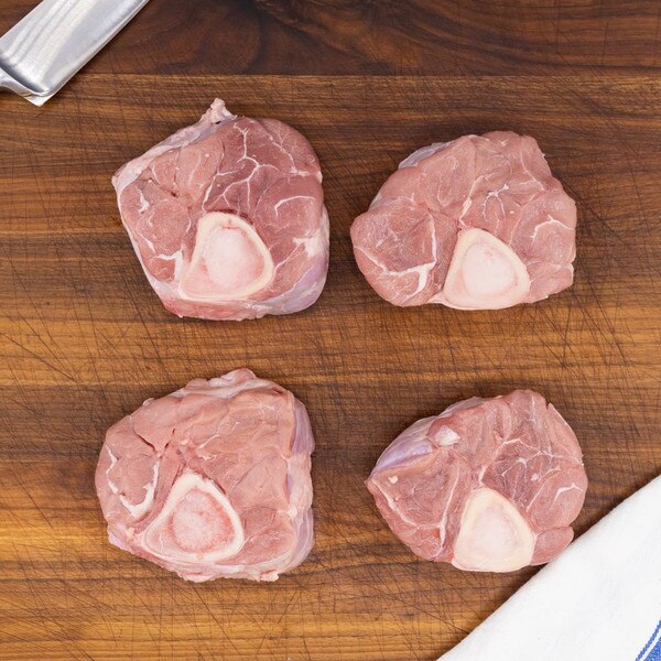 Four raw milk-fed French veal hindshanks crosscut for osso bucco on a wooden board with a white towel across the bottom right.
