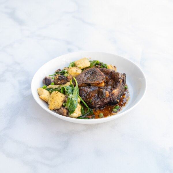 Tomato-braised milk-fed French veal osso bucco with Parisian gnocchi, English peas & spinach in a white bowl on a gray-and-white marble background.