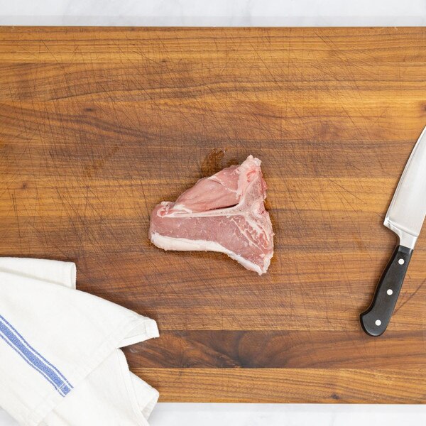 A raw, milk-fed French veal porterhouse chop on a wooden cutting board with a blue-striped, white towel on the left and a knife on the right.